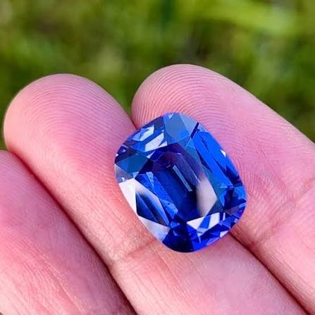 A vivid blue faceted tanzanite gemstone held between fingers against a blurred green natural background